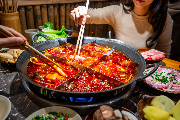 un bol de comida china cocinandose sobre fuego y una mujer con palillos chinos escogiendo uno de los platillos